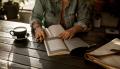 Person reading and writing notes at a table with books and a cup of coffee