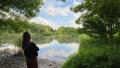 Person standing beside a peaceful lake surrounded by trees and natural greenery