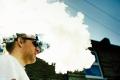 Person exhaling a large cloud of vapor outdoors, representing nicotine use, vaping, and substance-related behaviors