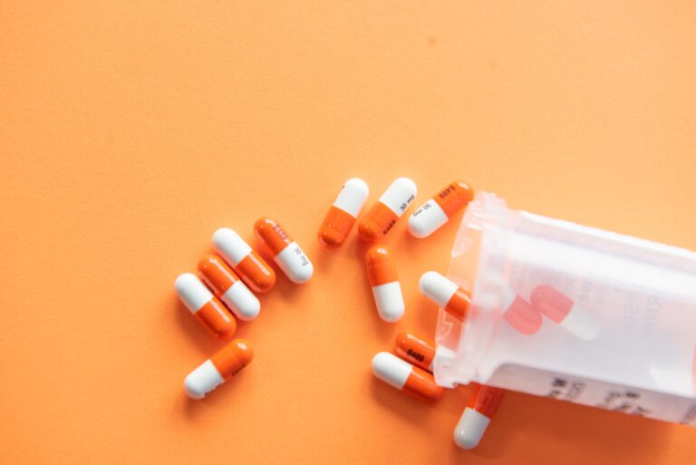 Orange and white prescription capsules spilling from a medication container onto an orange surface