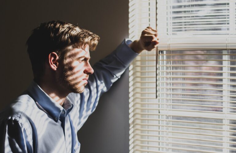 Man standing by a window looking outside, appearing thoughtful and reflective in natural sunlight