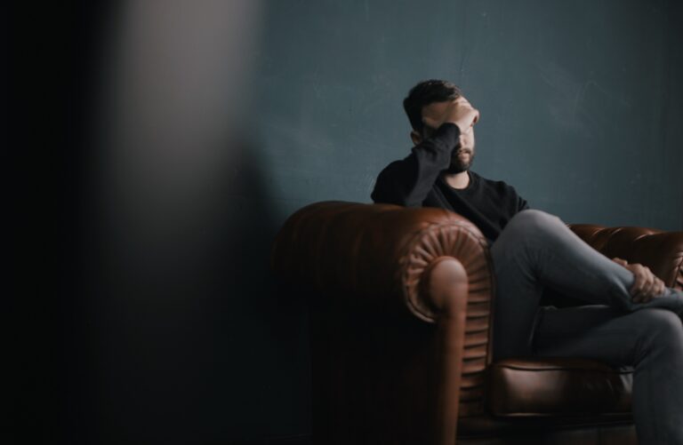Man sitting alone on a couch with his hand covering his face, reflecting emotional distress and mental health challenges