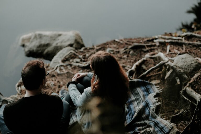 Two people sitting together by a lakeside surrounded by tree roots, reflecting and sharing a quiet moment of connection