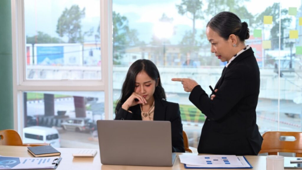 A distressed employee sitting at a desk while a supervisor points at them during a workplace interaction