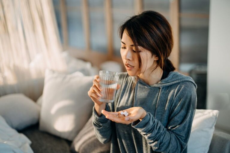 Woman holding sleeping pills and a glass of water while sitting at home, showing the stress of sleep deprivation and substance use