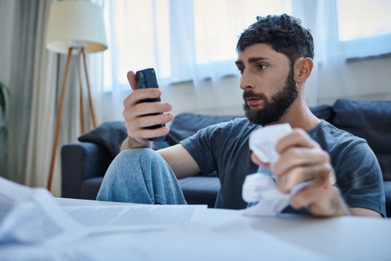 Stressed man looking at smartphone while holding bills after mobile gambling losses