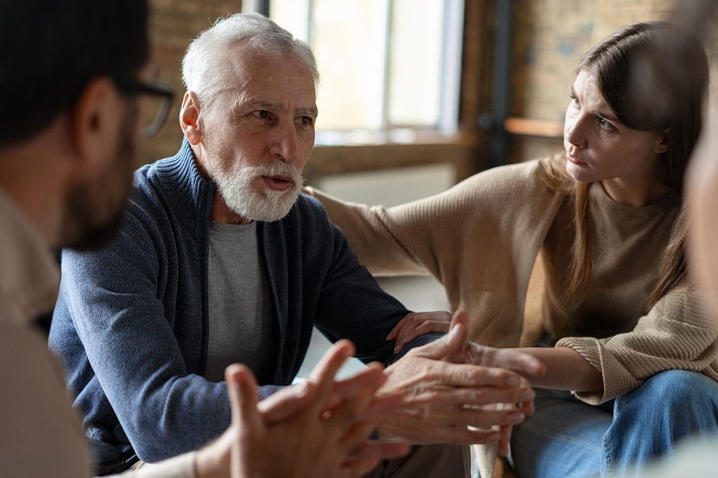 Older man speaking in a support group while a younger woman sits beside him, showing connection and support in addiction recovery